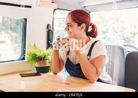 Junge Frau, die Kaffee trinkt und sich in ihrem handgemachten Wohnmobil entspannt. Roadtrip im Minibus und Sommerabenteuer im Freien. Nomad Lifestyle-Konzept Stockfoto