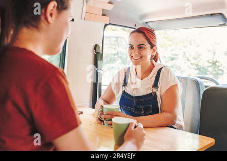 Zwei junge Frauen, die Kaffee trinken und sich in einem handgemachten Wohnmobil amüsieren. Roadtrip im Minibus und Sommerabenteuer im Freien. Nomad-Lebensstil Stockfoto