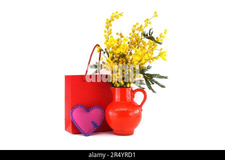 Strauß gelber Mimosablüten in roter Vase nahe roter Geschenktasche isoliert auf weißem Hintergrund, Ostern, 8. März Frauentageskonzept Stockfoto