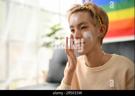 Ein glücklicher junger asiatischer Schwuler trägt Sonnencreme oder Feuchtigkeitscreme auf sein Gesicht auf, bevor er sich zu Hause schminkt. LGBTQ+ Lifestyle-Konzept Stockfoto