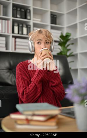 Portrait, ein attraktiver und entspannter junger asiatischer Schwuler, genießt seinen Kaffee, während er Musik über seine Kopfhörer im Wohnzimmer hört. Stockfoto