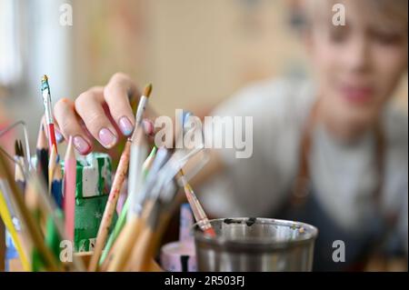 Nahaufnahme eines glücklichen, jungen asiatischen Schwulen, der Pinsel in einem Bleistifthalter auswählt und Malerei in einer Kunstwerkstatt genießt. Bild mit selektivem Fokus Stockfoto