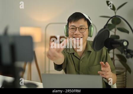 Ein fröhlicher Mann, der über Kopfhörer mit einem Kondensatormikrofon Podcast in einem kleinen Heimstudio aufnimmt Stockfoto