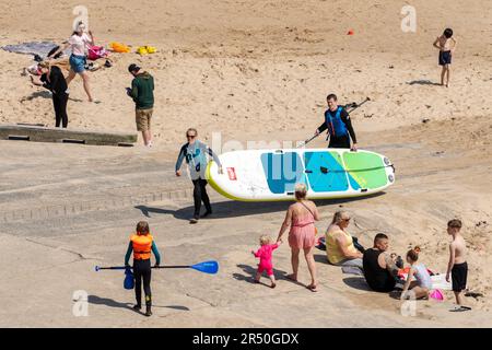 Menschen, die den Strand bei sonnigem Wetter genießen, einschließlich Stand-Up-Paddleboarder mit ihrem Brett. Cullercoats Bay, North Tyneside, Großbritannien Stockfoto