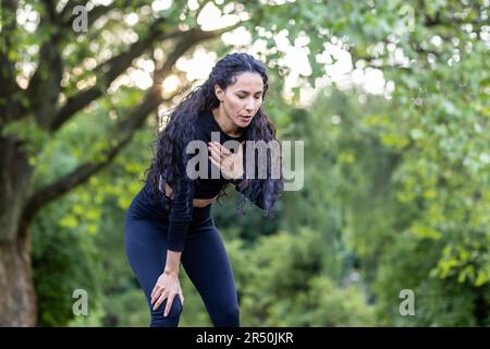 Porträt einer wunderschönen Sportlerin im Park, hispanische Frau ruht und atmet beim Joggen und aktiven körperlichen Training, sportliche Sportlerin hat Brustschmerzen. Stockfoto