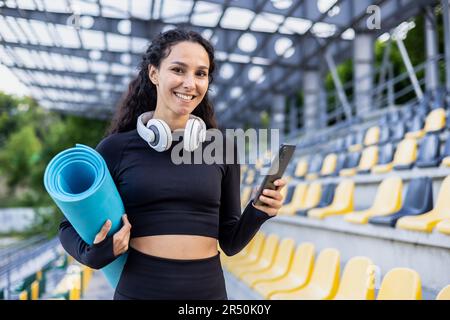 Porträt einer glücklichen Sportlerin, einer hispanischen Frau, die vor dem Stadion lächelt und in die Kamera schaut, das Telefon und die Trainingsmatte hält und die App für das Co-Training nutzt. Stockfoto