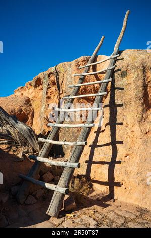 Vertikale Ansicht einer langen, verwitterten Pfahlleiter, die an einer sonnenbeschienenen orangefarbenen Tuffwand im Bandelier-Nationalmonument in New Mexico lehnt. Stockfoto
