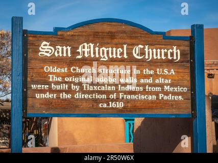 Ein blau gerahmtes Holzschild mit der Aufschrift San Miguel Church beschreibt das Adobe-Gebäude, mit einer Adobe-Mauer und blauem Himmel dahinter in Santa Fe, New Mexico. Stockfoto