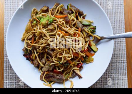 chinesische Nudeln mit Gemüse und Fleisch auf einem Holztisch mit natürlichem, sanftem Licht. Gebratene Nudeln in weißer Schüssel zu Hause auf dem Tisch umrühren. Plattiert und hausgemacht Stockfoto
