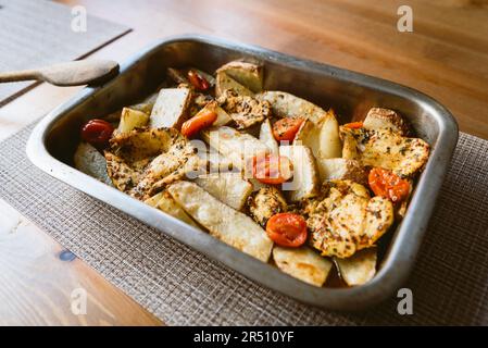 Hausgemachtes Hähnchen aus dem Ofen mit Kartoffeln und Tomaten auf einem Holztisch in einem Aluminiumtablett. Mariniertes Hähnchen, das für den sonntagsbraten im Ofen gebacken wurde Stockfoto