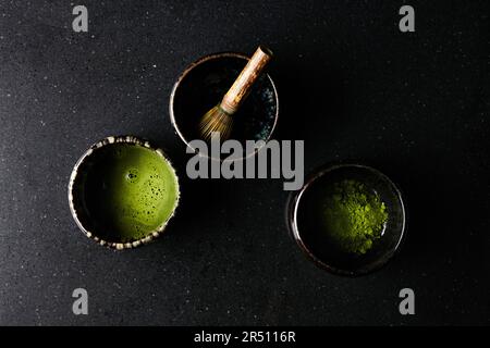 Matcha-Tee mit Matcha-Schneebesen und Teepulver in kleinen Schüsseln auf dunklem Hintergrund Stockfoto