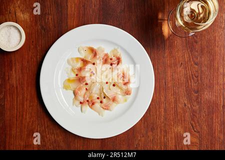 Fisch-Carpaccio mit roten Pfefferkörnern serviert mit einem Glas Weißwein Stockfoto