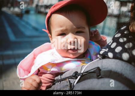 Nahaufnahme eines Babys, das während der Ferien von ihrer Mutter in einem Babyrucksack getragen wird. Das Reisekonzept für Kinder Stockfoto