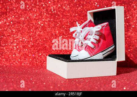 Ein Paar winzige rote Leinenschuhe in einer kleinen Geschenkbox, isoliert auf einem glitzernden roten Hintergrund. Hintergrund des elektronischen Handels und des Einkaufs. Stockfoto