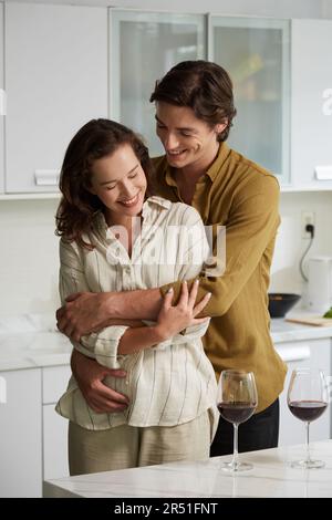 Glücklicher junger Mann, der verliebt ist und seine Freundin von hinten umarmt, wenn sie Wein in der Küche trinken Stockfoto