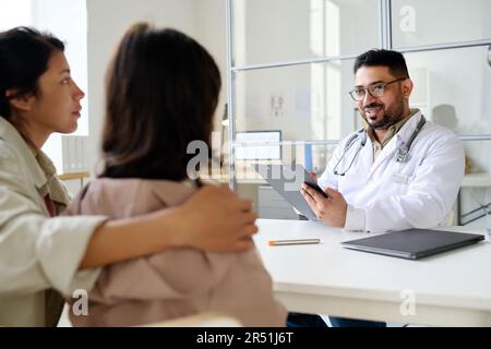 Kinderärztin, die mit der kleinen Patientin spricht, während sie ihm gegenüber sitzt, zusammen mit ihrer mutter während ihres Klinikbesuchs Stockfoto