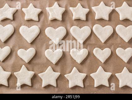 Rohe Kekse in Form von Sternen und Herzen auf einem Backblech mit Pergament, Draufsicht. Bühne der Zubereitung köstlicher hausgemachter Kuchen. Stockfoto