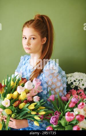 Porträt eines hübschen, jungen, 15 - 16 Jahre alten, rothaarigen Mädchens, umgeben von vielen Frühlingsblumen auf grünem Hintergrund, Schönheit und Modekonzept Stockfoto