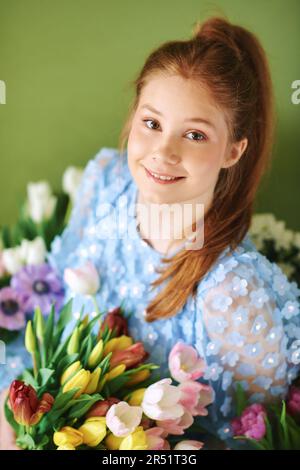 Porträt eines hübschen, jungen, 15 - 16 Jahre alten, rothaarigen Mädchens, umgeben von vielen Frühlingsblumen auf grünem Hintergrund, Schönheit und Modekonzept Stockfoto