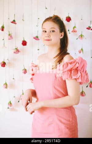 Studio-Porträt eines hübschen jungen Teenagers im Alter von 15 bis 16 Jahren, rothaarig, rosa Korallenkleid, Posen auf weißem Hintergrund mit hängenden Blumen, BE Stockfoto