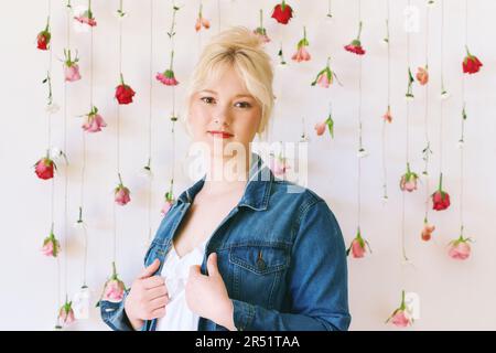 Studioporträt eines hübschen jungen Mädchens im Alter von 15 bis 16 Jahren, das eine Jeansjacke trägt und auf weißem Hintergrund mit hängenden Blumen, Schönheit und Mode posiert Stockfoto