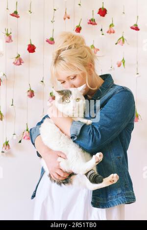 Studioporträt eines hübschen jungen Mädchens im Alter von 15 bis 16 Jahren, das eine Jeansjacke trägt, auf weißem Hintergrund mit hängenden Blumen posiert, eine süße Katze in der Hand hält, Stockfoto