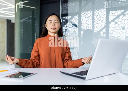 Junge, wunderschöne asiatische Webentwicklerin, die in einem Bürogebäude arbeitet, Frau, die mit geschlossenen Augen meditiert, Frau, die mit einem Laptop Code schreibt Stockfoto