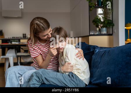 Der beleidigte Junge sitzt zu Hause mit seiner fürsorglichen Mutter auf der Couch. Lächelnde Mutter unterhält beleidigten kleinen Sohn Stockfoto