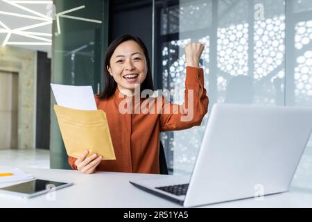 Porträt einer glücklichen Frau, asiatische Frau erhielt einen Brief, die glücklich und in die Kamera schaute, Geschäftsfrau, die in einem modernen Bürogebäude mit einem Laptop arbeitete. Stockfoto