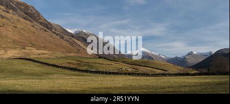 Glen Nevis Panorama vom Beginn des Ben Nevis Touristenpfads; sonnenbeleuchtete Felder im Vordergrund; schneebedeckte Hügel im Hintergrund; blauer Himmel mit weißen Wolken Stockfoto