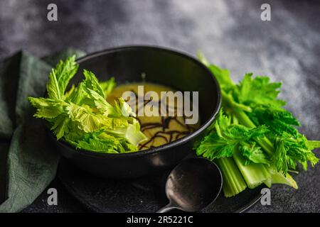 Vorderansicht einer gesunden Selleriesahne in einer Schüssel mit Selleriestangen serviert auf schwarzem Teller mit schwarzem Löffel und Serviette vor verschwommenem dunklem Hintergrund Stockfoto