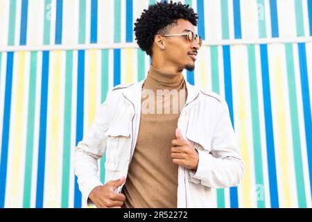 Ein glücklicher junger afroamerikanischer Mann mit Sonnenbrille und frischer Frisur, der seine Jacke in der Hand hält und wegschaut und an eine Wand lächelt Stockfoto