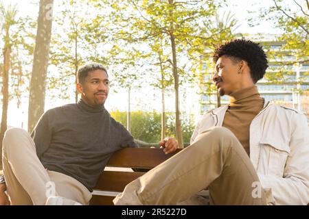 Junge afroamerikanische Freunde sitzen auf einer hölzernen Bank und schauen einander an, während sie sich während eines Meetings in einem Park unterhalten Stockfoto