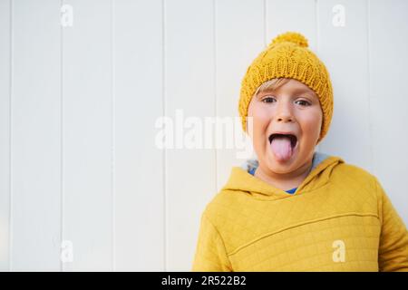 Nahaufnahme eines bezaubernden 5-jährigen Kindes mit gelbem Sweatshirt und Hut, Posen auf weißem Hintergrund, Zunge ziehen Stockfoto