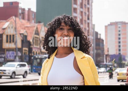 Positive afroamerikanische Frau mit lockigem Haar, die lächelt und in die Kamera schaut, während sie auf der Straße der Stadt vor verschwommenen Gebäuden und Autos steht Stockfoto