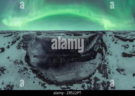 Weitwinkelblick aus der Vogelperspektive auf die majestätische Landschaft mit leuchtend grünen Polarlichtern über verschneiten Felsformationen in Island Stockfoto
