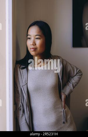 Porträt einer ernsten jungen asiatischen Frau in legerer Kleidung, die am Fenster steht und bei hellem Tageslicht wegschaut Stockfoto
