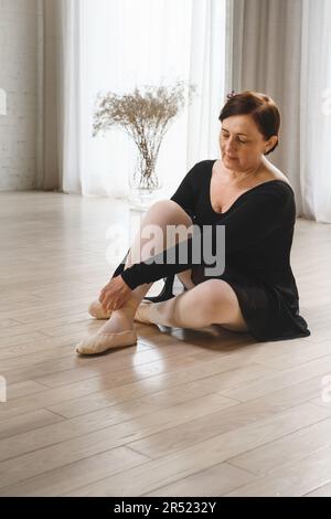 Der ganze Körper einer fokussierten, reifen Tänzerin in schwarzem Kleid sitzt auf dem Boden und zieht Ballett-pointe-Schuhe an, während sie sich auf die Probe im Studio vorbereitet Stockfoto