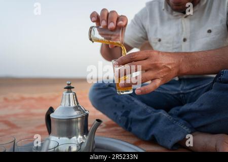 Vorderansicht eines verkürzten, anonymen afroamerikanischen Mannes in legerer Kleidung, der auf einem Teppich sitzt und erfrischenden heißen Tee einschenkt Stockfoto