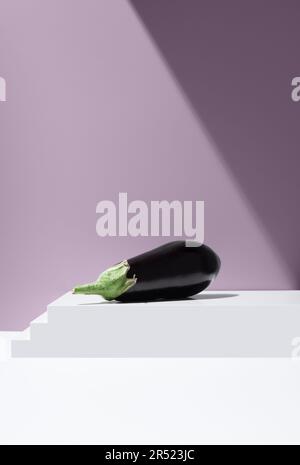 Lebhaftes Bild einer schwarzen Aubergine mit grüner Kappe auf einer weißen, gestuften Scheibe vor violettem Hintergrund bei hellem Licht Stockfoto
