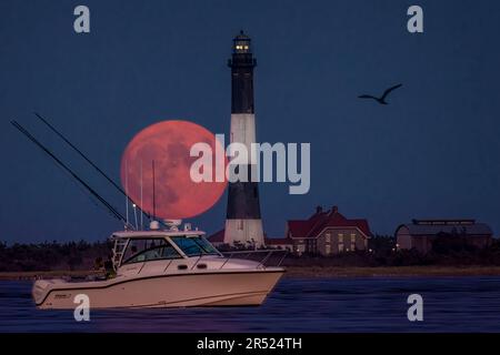 Fire Island Lighthouse Moon - der Vollerntmond erhebt sich hinter dem Fire Island Lighthouse und einem Freizeitboot in der Bucht. Fire Island Leuchtturm i Stockfoto