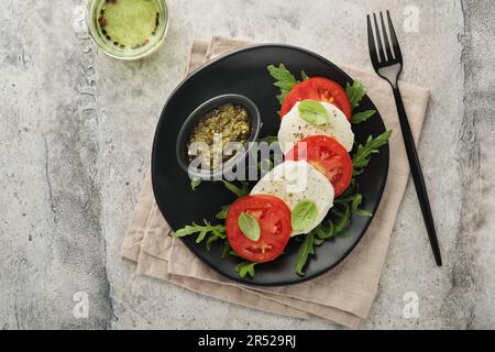 Caprese-Salat. Italienischer Caprese-Salat mit Tomaten, Mozzarella-Käse, Rucola, Basilikum, Olivenöl auf schwarzer Platte über altem Betongrauen Backgrou Stockfoto