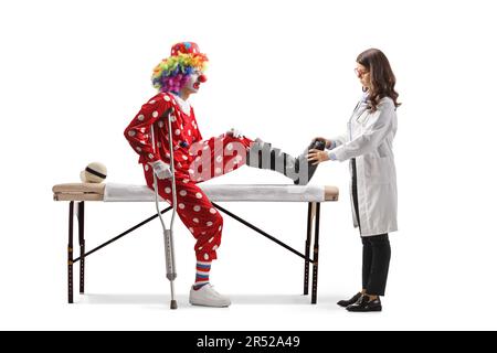 Verletzter Clown mit einer Wanderspange, der auf einem Therapiebett sitzt und der Arzt seinen Fuß isoliert auf weißem Hintergrund untersucht Stockfoto