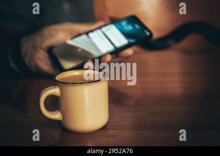 Von oben: Eine nicht wiedererkennbare ältere Person, die mit Nachrichten auf den Bildschirm des Mobiltelefons schaut, während sie mit einer Tasse heißem Kaffee am Tisch sitzt, gegen verschwommen Stockfoto