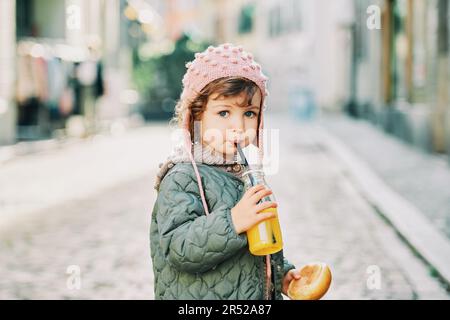 Außenporträt eines niedlichen Kleinkindes, das ein Milchbrotbrötchen hält und Orangensaft aus der Flasche trinkt Stockfoto