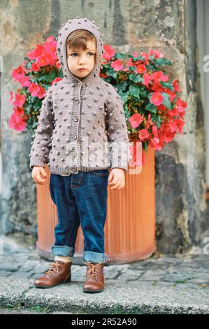 Außenporträt eines niedlichen Kleinkindes, das eine braune Strickjacke und dunkelblaue Jeans trägt, Mode für kleine Kinder Stockfoto