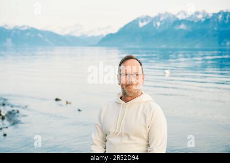 Portrait eines gutaussehenden Mannes, der einen wunderschönen See mit Bergen bewundert, mit weißem Sweatshirt Stockfoto