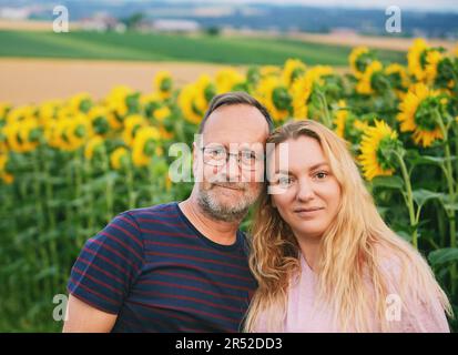 Außenporträt eines Paares mittleren Alters, das einen schönen Tag auf dem Land genießt und neben dem Sonnenblumenfeld posiert Stockfoto