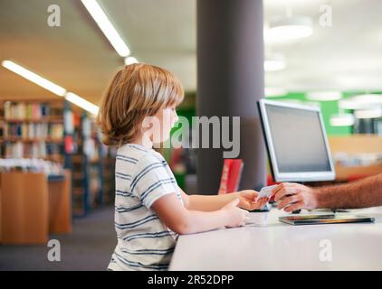 Porträt eines süßen kleinen Jungen, der Bücher in die Bibliothek nimmt und dem Bibliothekar einen Mitgliedskarten gibt Stockfoto