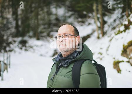 Außenporträt von 55 bis 60 Jahren, wandern im Winterwald, trägt warme Jacke und schwarzen Rucksack Stockfoto